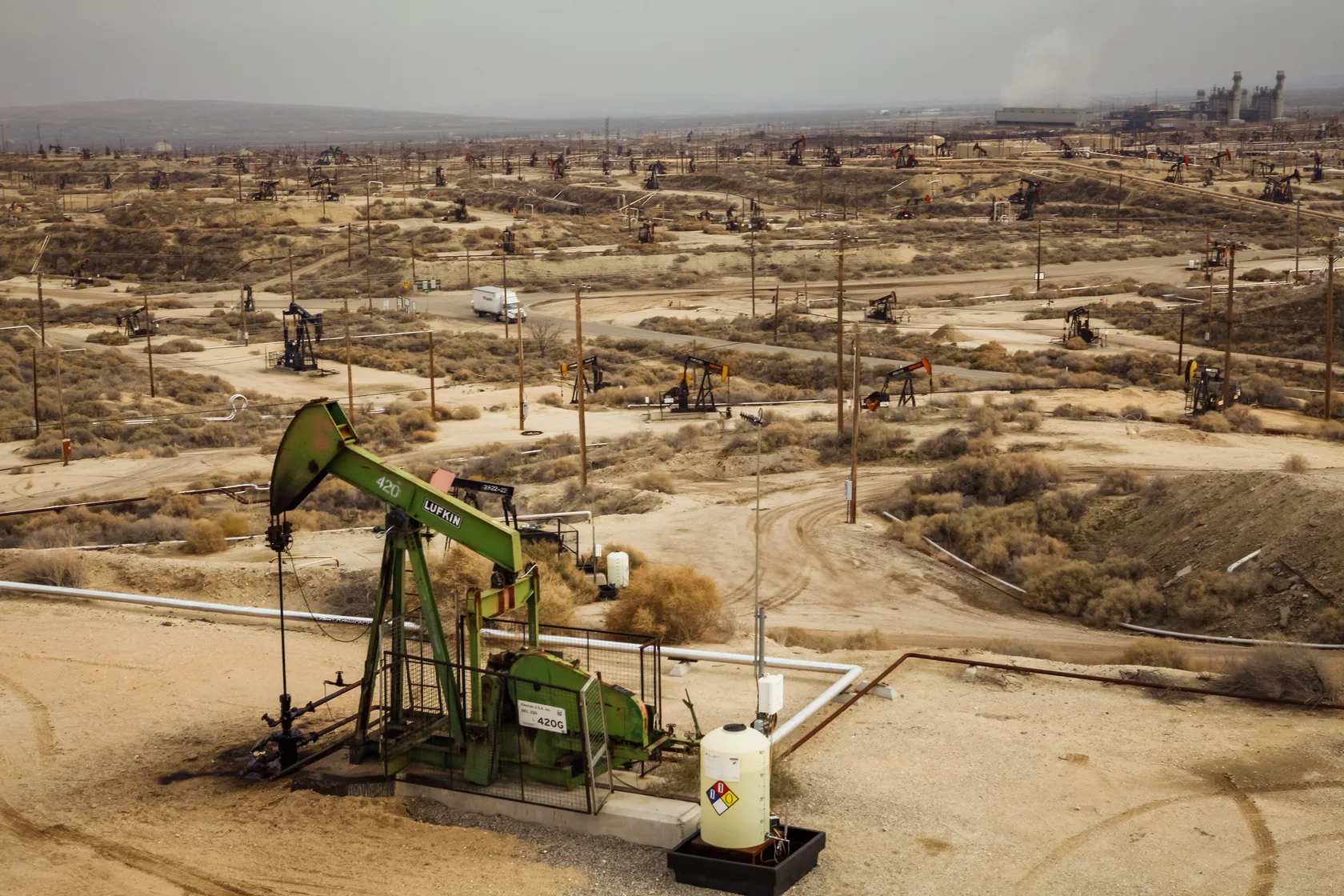 A green pumpjack in the foreground of a wide Kern County oil field dotted with well heads